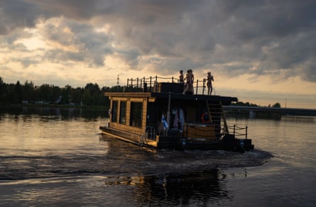 People in swimming costumes stand on the roof of a wooden cabin that floats on a lake