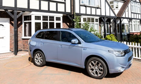 a silver-blue Mitsubishi Outlander PHEV, a large SUV, is seen side-on parked on a tiled driveway in front of a house with a mock-Tudor black and white frontage, white front door and windowframes, and white picket fence. A yellow cable is seen emerging from the charging port which is behind a flap in the car's rear wing, and this trails behind the car to the power source.