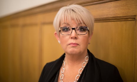 In front o f a wooden panel Helen Newlove, with short hair, glasses, a necklace with big links and a jacket, raises her eyebrows