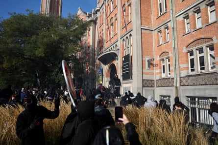 A crowd of people, who appear to be mostly male and wearing black or grey hoodies, moves towards the doorway of the redbrick town hall. Some are holding up phones to film the entry, and one person facing the camera wears a ski mask to obscure their face.