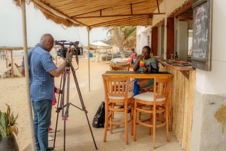 An African woman sits at a beachfront cafe sipping a drink while being filmed by a man
