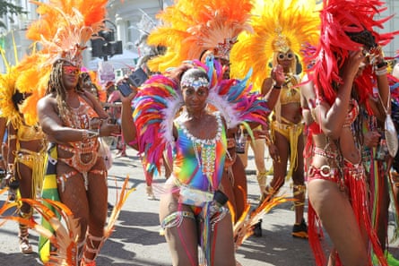 a group of women in revealing but elaborately decorated and brightly coloured costumes and huge feathered headdresses dance in the street