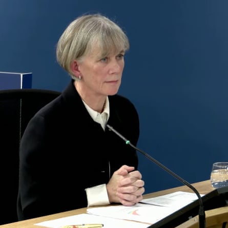 Angela McLean, with short hair and a button down shirt under a blazer, sits with her hands folded in front of a microphone