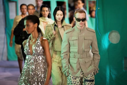 A line of models. The woman at the front wears a silver sequinned halterneck dress; behind her two women wear military-style jackets and trousers