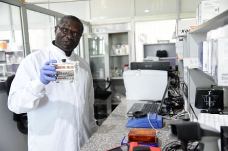 A man in a white lab coat and purple nitrile gloves holds up a piece of equipment in a laboratory.