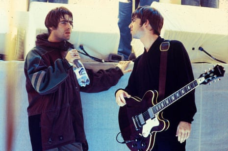 Ο Noel Gallagher κρατά μια κιθάρα και στρίβει δεξιά για να μιλήσει στον Liam Gallagher που κρατά ένα μπουκάλι νερό