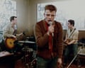 Suggs performs into a microphone while two bandmates play guitars, from the film Take It or Leave It