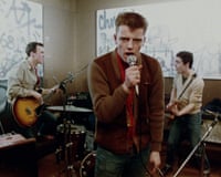 Suggs performs into a microphone while two bandmates play guitars, from the film Take It or Leave It