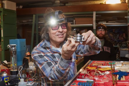 A man in a checkered shirt and a head lamp tinkers on a project in a tool shed with engineering supplies