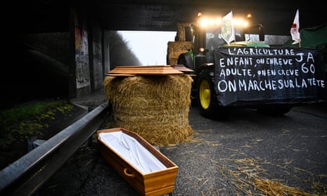 A French farmer rides his tractor with a banner reading 'Farming, as a child we dream of it, as an adult we die of it, we are walking on our heads' next to a coffin, as they block the A16 highway.