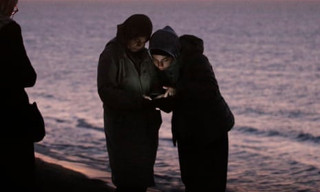 The faces of two young women wearing head coverings and jackets, standing very close to a shore that reflects dark blue and pink light from a nearly set sun, are illuminated by a phone they hold between them.