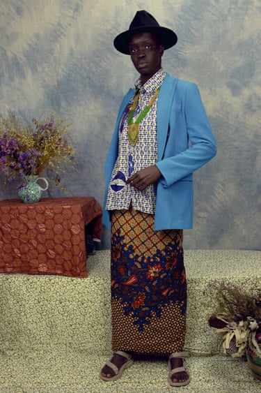 The artist dressed as a man in a black hat and blue blazer wears a patterned shirt and long printed skirt