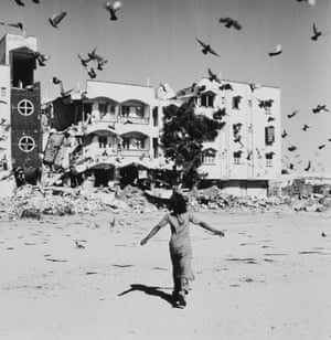 A black and white picture shows a child running after birds that fly into the air. In the background is a badly damaged building.