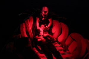 A torch shows a rescued young man lying with eyes closed on the floor of a small boat, his head cradled by a rescue worker