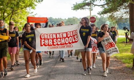 On a rural-looking asphalt lane between two grassy verges and beneath leafy trees, a group of mixed race young people wearing black T-shirts that says 'Sunrise Movement' in yellow lettering, walk in a group holding signs, with the two girls in front holding a white banner that says in red lettering 'Green New Deals for Schools, Summer Camp 2023.'