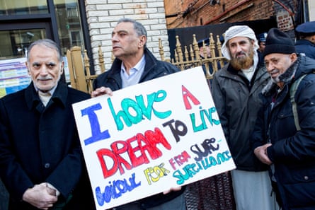 Four middle-aged men who might be Arab stand in black winter coats on a city street, with one holding a multicolored sign.