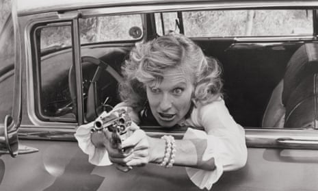 Cloris Leachman leaning out of the driver's side of a 1950s car with a wild expression, pointing a large revolver at somebody offscreen