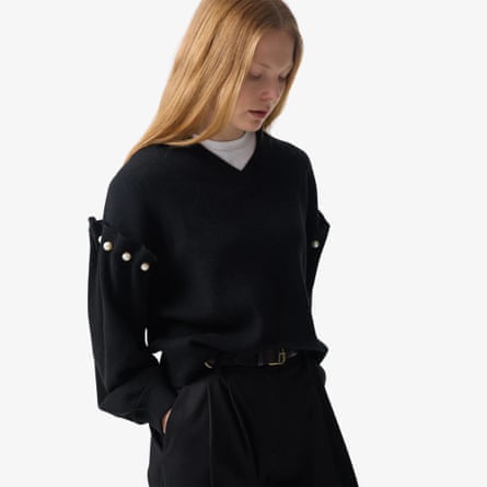 A model with long blond hair wears the Tallie jumper in black with pearl details. They have their hands in pocket and look downwards. The model also wears a white T-shirt under the jumper an black tailored trousers with a belt