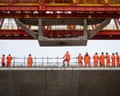 Workers in orange he-vis and hard hats look up at concrete section being moved by machinery