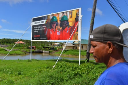 Man walks past a billboard in a rural setting put up by ExxonMobil extolling the opportunities and benefits of oil and gas exploration to Guyana.