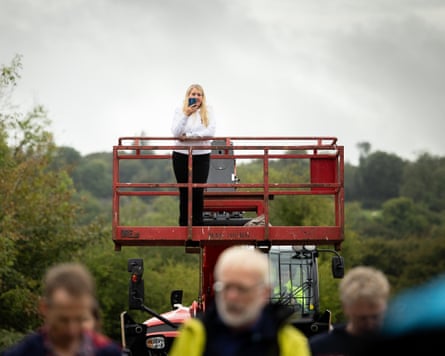 A woman stands on a red cherrypicker filming a road bowling event.
