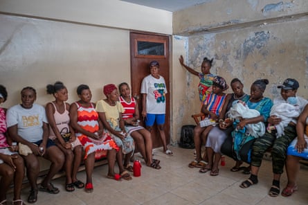 Women, some holding babies, sit or stand in a sparse room