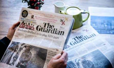 A person holding up the Guardian to read it, shot from over their shoulder; in the background is a table with a pot of tea and another copy of the paper on it