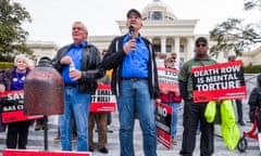 White man wearing hat, blue buttondown and jeans holds microphone while other people stand behind him holding signs.