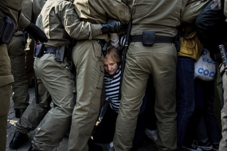 A protester on their knees is blocked by a line of police officers in green uniforms