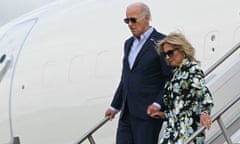 Older white man wearing suit without tie and aviator sunglasses, holds hand of older blond white woman wearing dark sunglasses and a floral, long-sleeved dress as they walk down steps out of a plane.