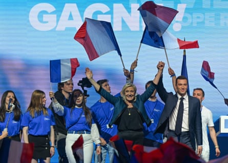 Marine Le Pen and Jordan Bardella wave French flags on a stage.