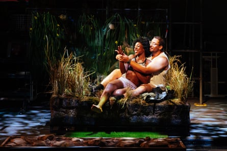 A male actor with his arms wrapped around a female actor as they are both sat on a dimly lit stage decorated with foliage.