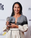 Monica Ali: she is seen wearing a grey, black and white dress with long gathered sleeves and skirt, holding an award. She is in her mid-50s; her long, straight dark hair is worn loose and she is wearing dangling earrings. 