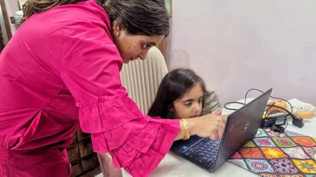A woman stands over a young girl working at a computer at a table.