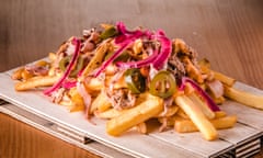 A wooden board on which sit loaded fries with pulled pork, jalapeños, cheddar sauce and onion.