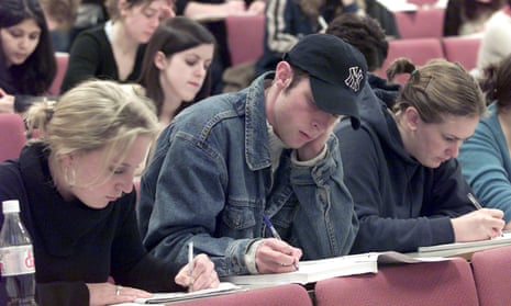 students taking notes in a lecture – close-up of three students with pens and notepads, two young women, one wearing a black polo neck jumper and the other a dark grey hoodie, both with their hair in ponytails, with a young man in denim jacket, cream jumper and baseball cap between them, all looking intent and concentrating