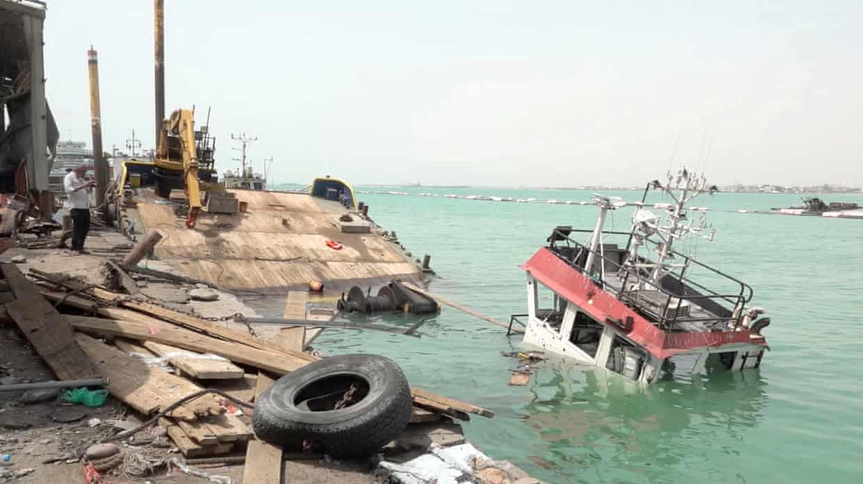 The control tower of a part-sunken boat appears above greenish water by a damaged wooden quayside with broken decking.