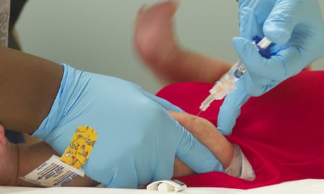 A close-up shot of a medical worker wearing blue gloves injecting the contents of a syringe into an infant's upper leg.
