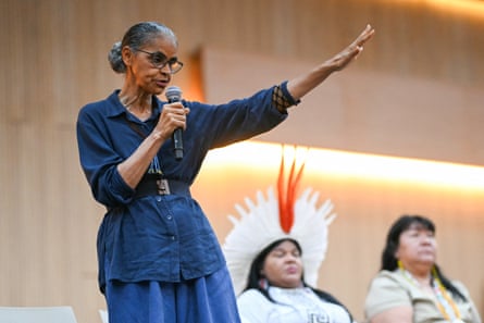 A woman with a microphone stands up with two women sit behind her, one in a traditional headdress