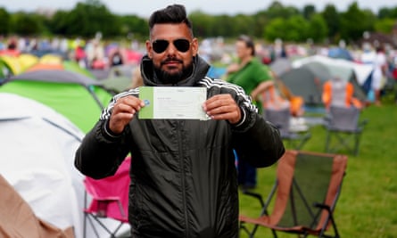 Blase, 41, holds up his number 1 queue ticket proudly: he has dark hair shaven up the side and gelled into a quiff on top, and a neat dark beard, and wears a black Adidas sports jacket with white stripes on the shoulders and Ray-Ban aviator sunglasses. He is standing in front of some camping chairs with tents seen in the background.