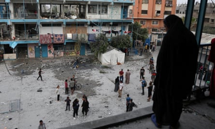 A woman dressed in black with a hood looks down at people standing in the space between school buildings. Rubble is on the ground and there are wrecked rooms in the building opposite