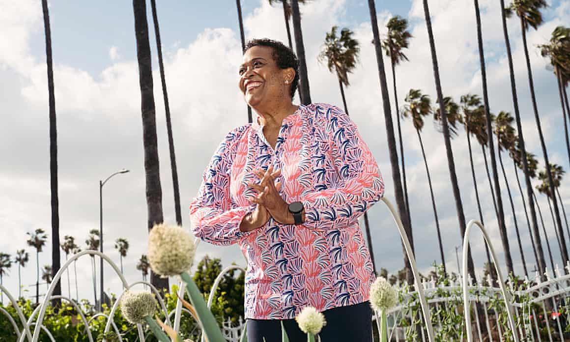 Beverly Lofton, in a bright pink top, smiles and clutches her hands in her garden in the View Park neighborhood of Los Angeles, a row of palm trees and blue sky behind her.