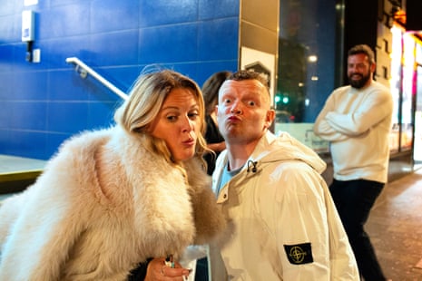 A woman and a man, both dressed in white, pouting their lips while a man in a cream jumper behind them is laughing
