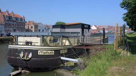 A black barge is moored on a canal bank with buildings and blue skies behind