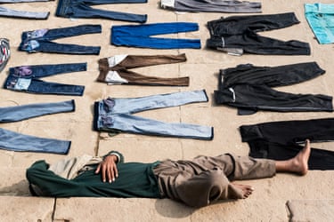 A man lies on stone ground amid rows of trousers laid out flat to dry