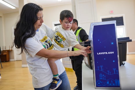 Woman holding child with facepaint of a rocket puts ballot in ballot box.