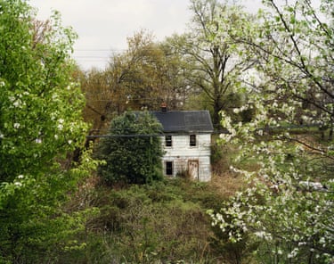 An attractive wooden cottage, painted white, in a wooded glade overgrown with bushes. Its front door, white with three small panes in a classic diagonal row, still looks welcoming