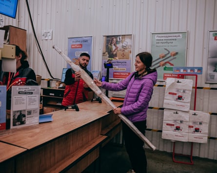 A woman holds a long tubular package standing at the counter in a post office with a clerk serving her