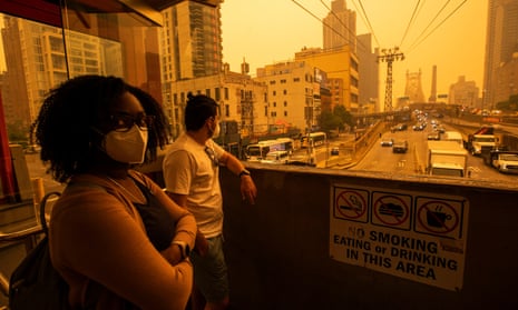 A young Black woman with curly hair, glasses, and a mask, beside a white or Latino man in white shirt, jeans and a mask, look out from an elevated station at the street below at a completely yellow sky.