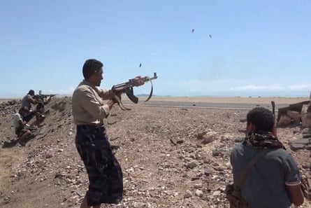 Fighters fire machine guns across a flat stretch of sandy, rocky land and road. Several point their guns from behind a low ridge of rocks and sand. They wear civilian T-shirts and shirts and several are bare-headed.